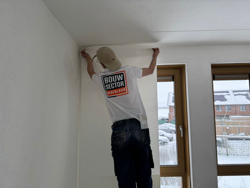 Behanger van Bouwsector Nederland plakt een baan renovlies bovenaan de wand bij het plafond
