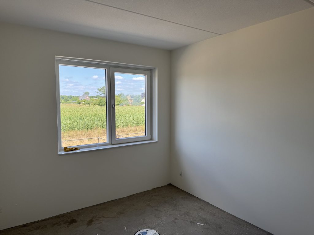 Lege slaapkamer in een nieuwbouwhuis met strak wit gesausde wanden en uitzicht op een maisveld