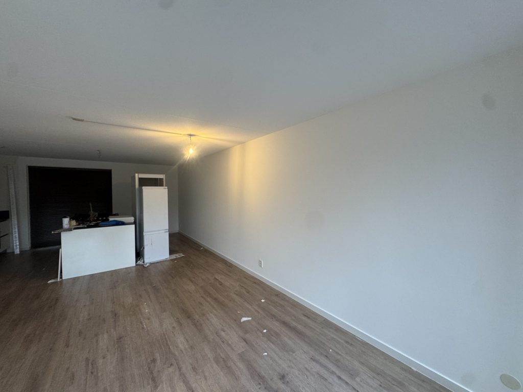 Woonkamer van een appartement met open keuken en een lange, strak gesausde witte wand